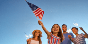 Happy family celebrating Independance Day. Young girl in the forefront waving a small American flag with mom, dad and brother in the background waving sparklers